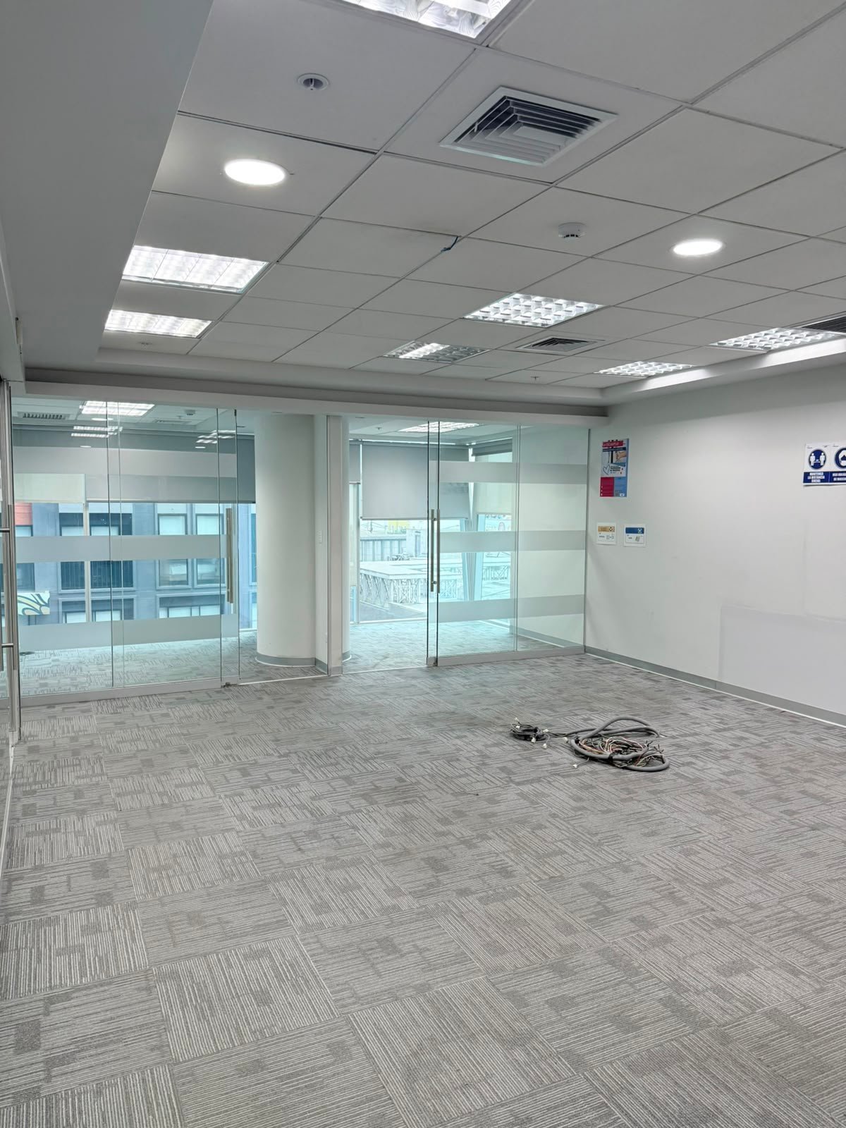 Empty modern office space with glass partition walls, fluorescent ceiling lights, beige tile flooring, and HVAC vent visible