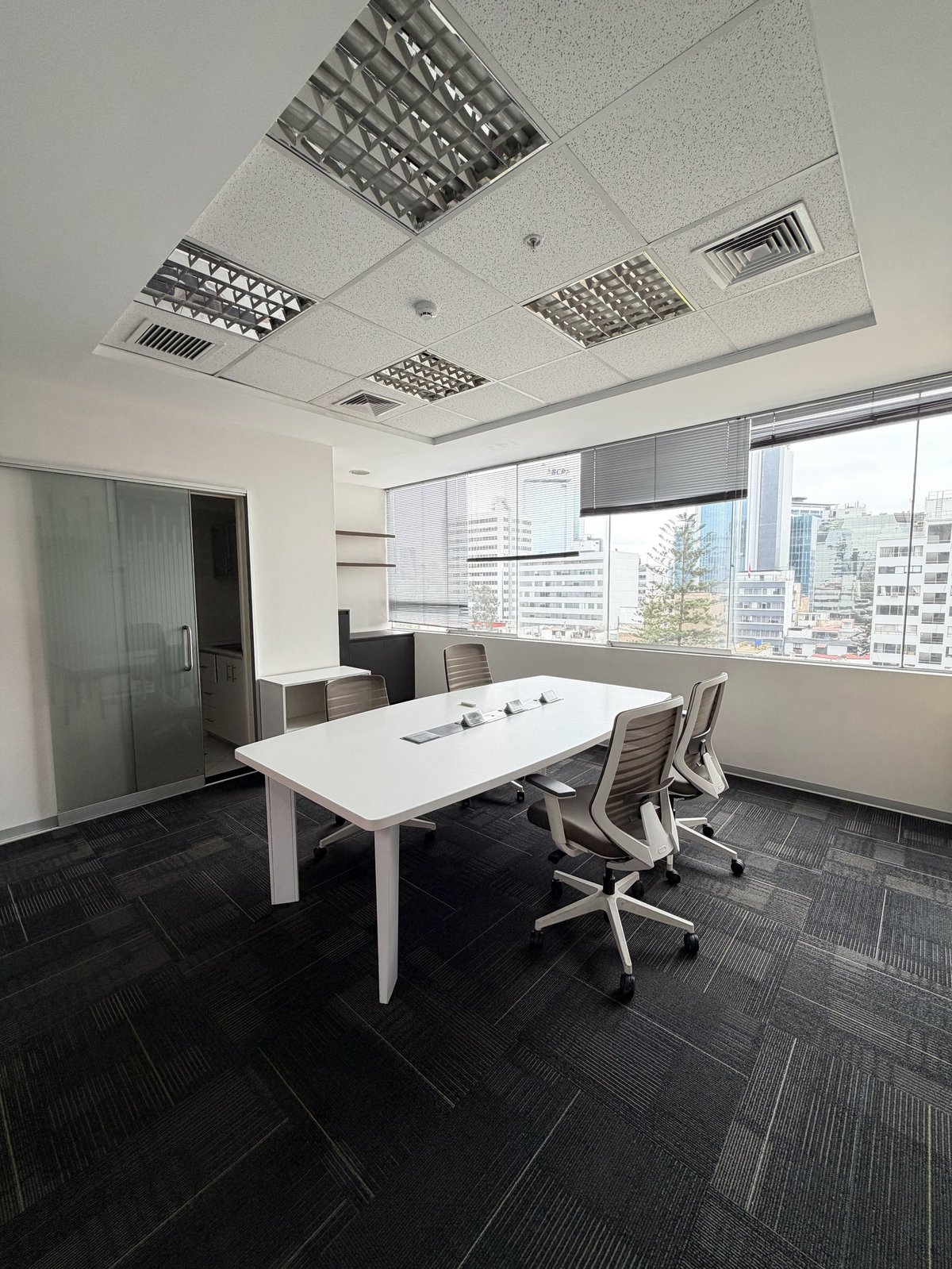 Modern office conference room with white table, ergonomic chairs, floor-to-ceiling windows overlooking city skyline, and recessed ceiling lights