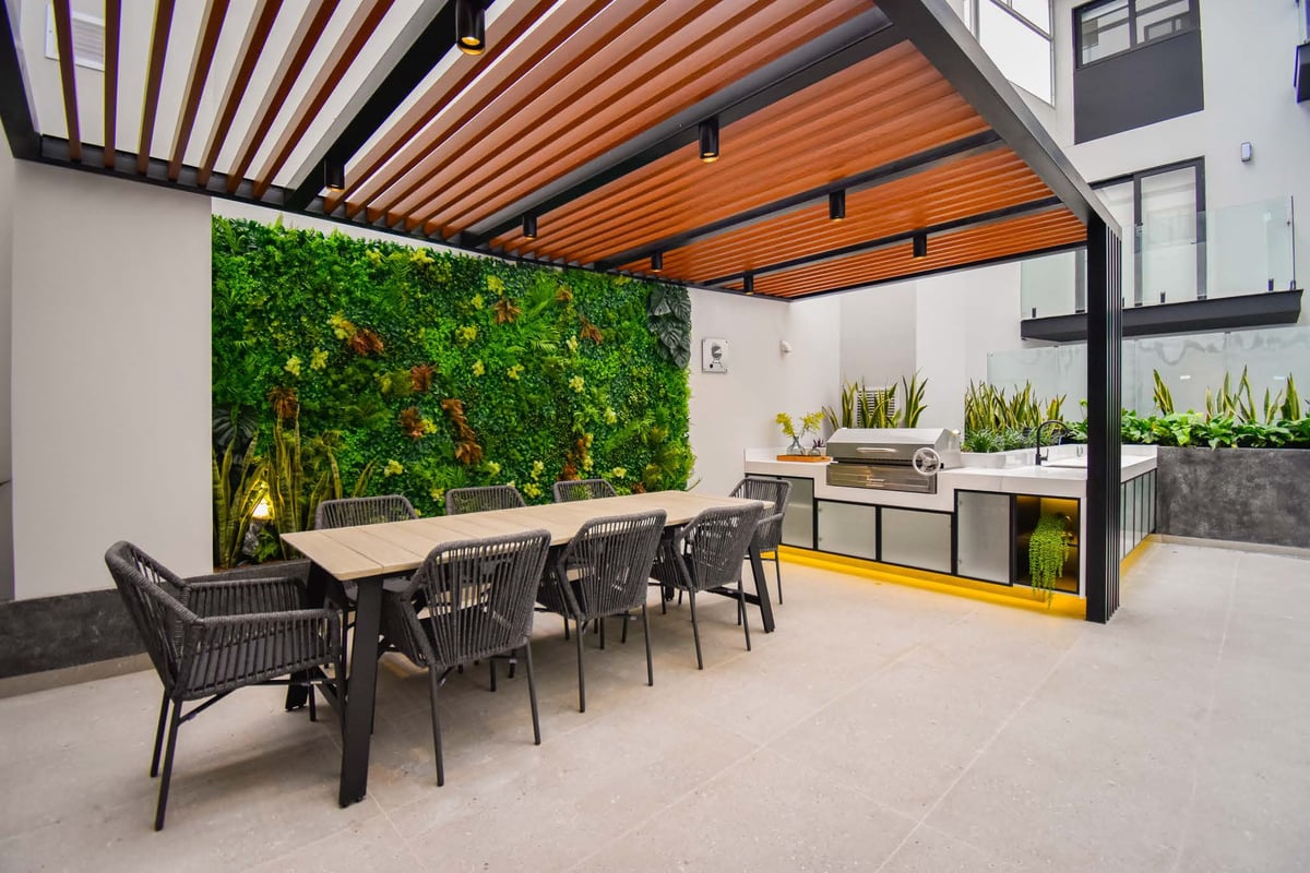 Modern outdoor dining patio with wooden pergola, living green wall, dining table with black chairs, and kitchen counter area